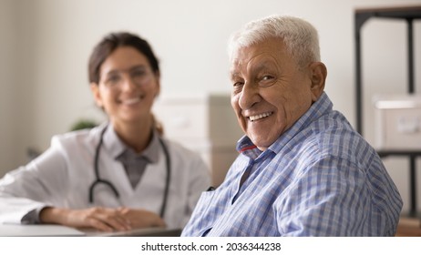 Happy Optimistic Senior 80s Man Looking At Camera With Toothy Smile During Appointment At Doctor Office. Portrait Of Elder Male Patient And Young Female GP Therapist In Background. Medic Care