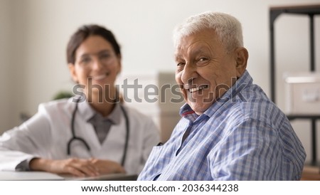 Happy optimistic senior 80s man looking at camera with toothy smile during appointment at doctor office. Portrait of elder male patient and young female GP therapist in background. Medic care