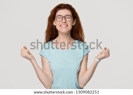 Happy overjoyed red-haired woman in glasses celebrating win success with yes gesture, excited female winner rejoicing victory new opportunity triumph isolated on blank grey white studio background
