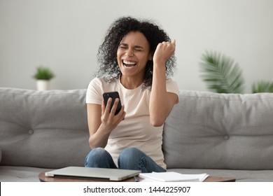 Happy Overjoyed Young African American Woman Holding Mobile Phone Celebrating Bid Win Game App Victory Sit On Sofa At Home, Excited Euphoric Black Girl Student Winner Rejoicing Success With Cell Phone