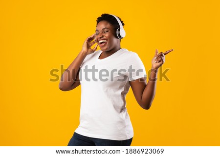 Happy Overweight Black Lady Wearing Headphones Enjoying Music Listening To Favorite Song Online Standing In Studio Over Yellow Background. Great Playlist Concept