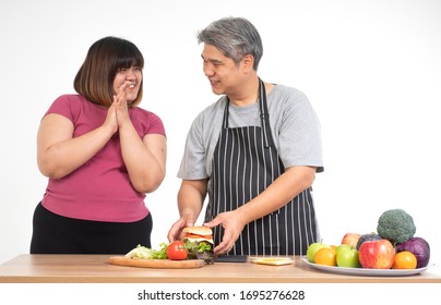 Happy Overweight Couple Cooking A Burger In The Kitchen Room. A Vegetable On The Table. Concept Of Happy Family And Binge Eating