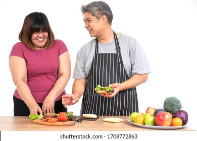 Happy Overweight Couple Cooking A Burger In The Kitchen Room. A Vegetable On The Table. Concept Of Happy Family And Binge Eating