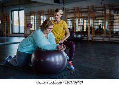 Happy Overweight Woman Exercising With Personal Trainer On Fintess Ball In Gym