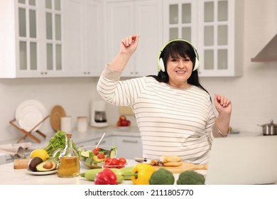 Happy Overweight Woman With Headphones Dancing While Cooking In Kitchen