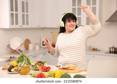 Happy Overweight Woman With Headphones Dancing While Cooking In Kitchen