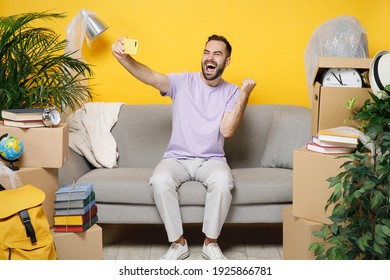 Happy owner man doing winner gesture doing selfie shot on mobile phone sit in living room on sofa at home unpacking stuff rent flat isolated on yellow wall. Relocation moving in apartment concept - Shutterstock ID 1925866781