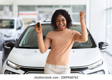 Happy Owner. Overjoyed Black Lady Holding Keys Of Her New Car After Buying, Cheerful African American Woman Standing Near Vehicle In Dealership Center And Raising Fist With Excitement, Copy Space