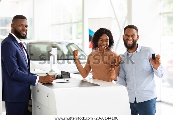 Happy Owners. Excited Black Couple Celebrating\
Buying New Car In Dealership Center, Cheerful African American\
Spouses Standing Next To Sales Desk, Holding Keys And Exclaiming\
With Joy, Copy Space