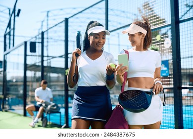 Happy paddle tennis player and her female friend using mobile phone on outdoor court. - Powered by Shutterstock