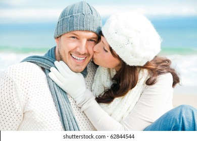 Happy Pair Of Male And Female Embracing And Having Fun Wearing Warm Clothes Outside On Coast Behind Blue Sky