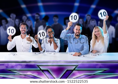 Happy Panel Judges Sitting In Front Of Red Curtain Showing 10 Score Signs