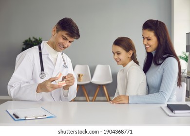 Happy Parent And Child Seeing Pediatric Dentist. Oral Hygienist With Jaw Model Teaching Patients To Brush Teeth Properly To Prevent Caries, Keep Teeth Clean And Healthy And Have Good Beautiful Smile