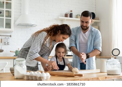 Happy parents and adorable little daughter cooking dough together, standing in modern kitchen, young mother and father teaching little girl to bake biscuits or muffins, family enjoying leisure time