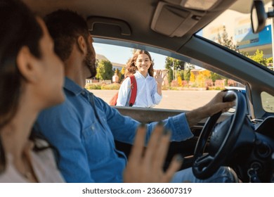 Happy parents bringing their daughter to school, sitting in car and waving hands to child girl. Mother and father saying goodbye to kid, before entering the school from the car - Powered by Shutterstock