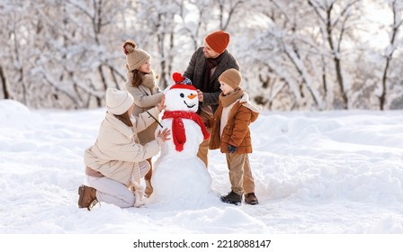 Happy parents and children gathering in snow-covered park together sculpting funny snowman from  snow. Father, mother and two kids playing outdoor in winter forest. Family active holiday