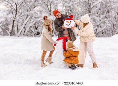 Happy parents and children gathering in snow-covered park together sculpting funny snowman from freshly fallen snow. Father, mother and two kids playing outdoor in winter forest. Family active holiday