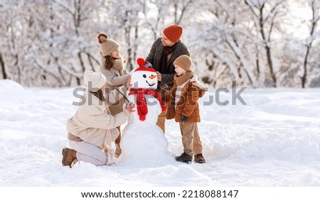 Happy parents and children gathering in snow-covered park together sculpting funny snowman from  snow. Father, mother and two kids playing outdoor in winter forest. Family active holiday
