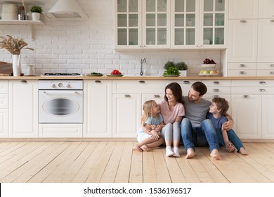 Happy parents with children sitting on warm wooden floor in modern kitchen, smiling father and beautiful mother hugging little cute daughter and son, family enjoying weekend at home together