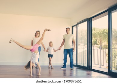 Happy parents and daughters celebrating housewarming, girl doing cartwheel. Full length. Apartment buying concept