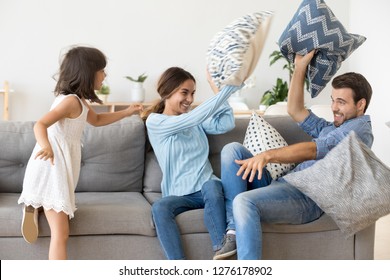 Happy Parents With Kid Daughter Having Fun Pillow Fight Enjoying Funny Activity On Weekend At Home, Cheerful Family And Little Child Girl Playing Laughing Together Throwing Cushions In Living Room