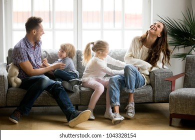 Happy Parents And Kids Having Fun Tickling Sitting Together On Sofa, Cheerful Couple Laughing Playing Game With Little Active Children Son And Daughter In Living Room At Home, Family Funny Activity