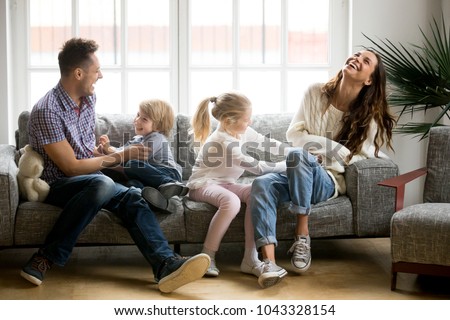 Happy parents and kids having fun tickling sitting together on sofa, cheerful couple laughing playing game with little active children son and daughter in living room at home, family funny activity