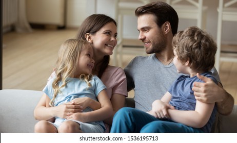 Happy parents with little daughter and son sitting on couch, looking at each other, enjoying tender moment together, children sitting on smiling mum and dad lap, cuddling, expressing love and unity