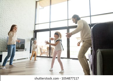 Happy parents play hide and seek with kids in big luxury country house with glass wall, blindfolded girl having fun with mom dad and little brother at home in living room, family active game concept