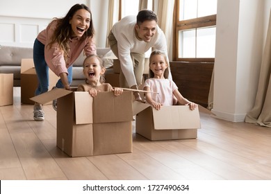 Happy parents playing with children in new apartment living room, two cute little daughters sitting in cardboard boxes, laughing mother and father pushing, family celebrating moving day