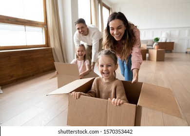 Happy parents playing with cute small kids daughters laughing on moving day, family tenants renters homeowners and children girls having fun riding in box in living room relocating new home concept