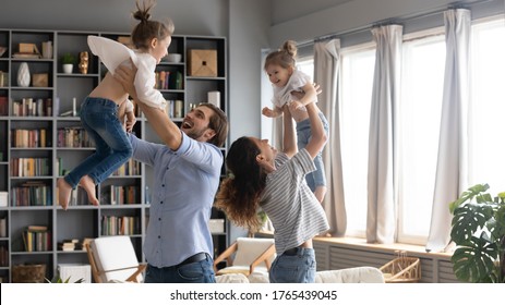 Happy Parents Playing Funny Active Game With Two Little Daughters, Standing In Living Room At Home, Smiling Mother And Father Lifting Cute Girls Pretending Flying, Enjoying Leisure Time Together