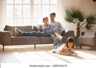 Happy parents relaxing couch in comfort light living room while little kid child daughter playing warm floor drawing and colored pencils  family having fun together  underfloor heating concept