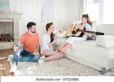 Happy parents sitting on carpet and looking at daughter playing guitar - Shutterstock ID 597253880