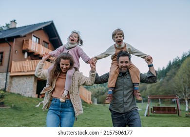 Happy Parents With Small Children On Piggybacks Running And Having Fun Together In Garden Near Their House.