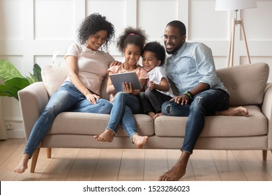Happy parents with smiling kids sitting on sofa, using tablet looking at screen, watching cartoons online, funny videos together, little girl showing family photos on gadget, making order, ecommerce