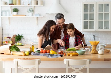 Happy Parents Teaching Kid Chopping Vegetables. Mom, Dad, Son Family Preparing Dinner Using Recipe In Cookbook. Cozy Home Interior. Positive Nice Communication And Warm Loving Relationship