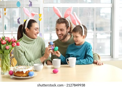Happy parents with their little son in bunny ears breaking Easter eggs at table