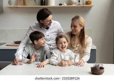 Happy parents and two healthy sibling kids drinking pure fresh clean water in kitchen, satisfying thirst, standing at table with glasses, smiling, laughing. Family care, children healthcare concept