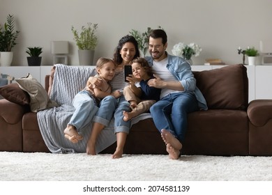Happy parents with two kids using smartphone at home together, sitting on cozy couch, smiling mother and father with little son and daughter looking at phone screen, taking selfie, having fun - Shutterstock ID 2074581139