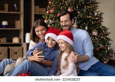 Happy parents and two little kids in Santa hats celebrating Christmas at glowing tree, taking festive selfie together, making video call for distance congratulations. Family holidays, communication - Powered by Shutterstock