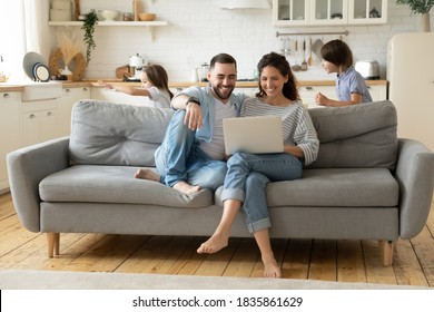 Happy parents using laptop, sitting on couch while kids playing funny game, smiling mother and father shopping online, enjoying leisure time, little son and daughter running and laughing together
