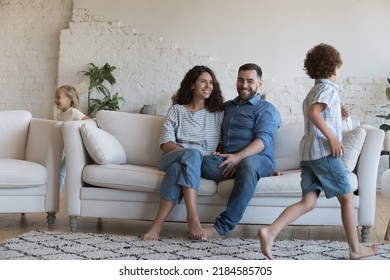 Happy Parents Watching Two Hyperactive Energetic Little Sibling Kids Running Around Sofa, Chasing, Playing Active Games In Living Room. Couple And Children Enjoying Family Leisure, Playtime