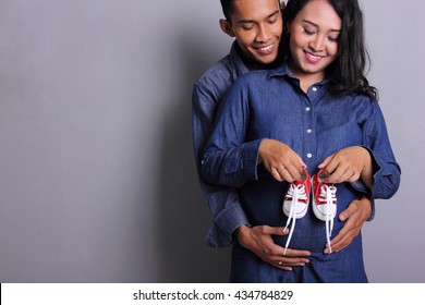 Happy parents-to-be couple looking at a cute red baby shoes for their unborn child, indoors studio portrait
