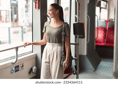 Happy Passenger Lady Standing In Modern Tram Enjoying Comfortable Ride In Public Transport Indoor, Looking Away Out Of Window. Student Commuting To University By Bus Or Trolleybus