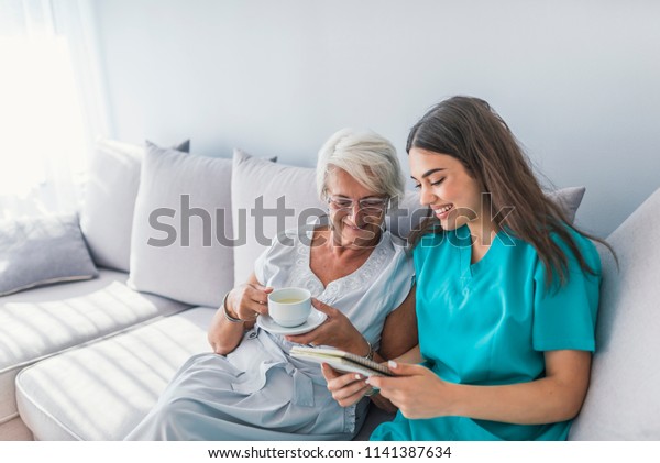 Happy patient is\
holding caregiver for a hand while spending time together. Elderly\
woman in nursing home and nurse. Aged elegant woman and tea time at\
nursing home
