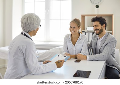 Happy Patients Talking To Family Doctor. Married Couple Planning To Have Baby And Seeing Gynecologist Or Pregnancy Specialist. Young Lady With Husband Or Boyfriend Sitting At Table At OB GYN's Office