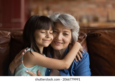 Happy Peaceful Loving Grandchild And Grandma Hugging Cheek To Cheek. Grandmother Meeting And Embracing Little Granddaughter With Love, Affection, Enjoying Time With Kid At Home. Family Bonding