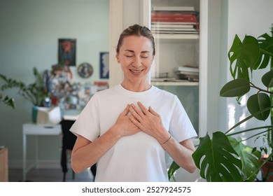 Happy peaceful middle aged woman standing alone indoor with closed eyes keep hands close to heart with hope pray in mind. Emotional female smiling feeling pleasure in deep gratitude. Thank you life. - Powered by Shutterstock