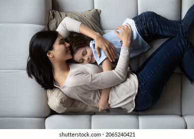 Happy peaceful young Asian mom hugging cute 7s son boy, sleeping on comfy sofa in living room. Calm sleepy mother and kid resting on soft couch, enjoying leisure time, recreation together. Top view